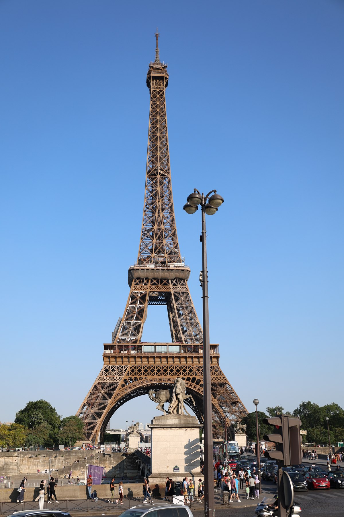 Photograph of the Eiffel Tower under a clear blue sky, with a double-headed street lamp crossing the frame in the foreground.